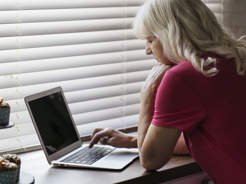 Mujer madura que usa la computadora portatil cerca de la ventana
